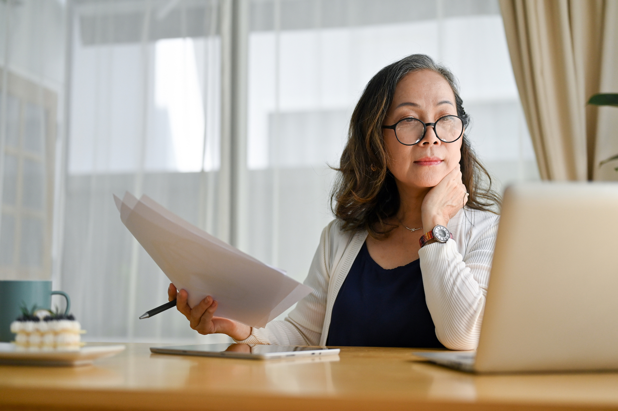 Concentrated asian middle aged female in glasses sitting at desk using portable computer and examining paperwork. How to Pay For Walk-In Tub